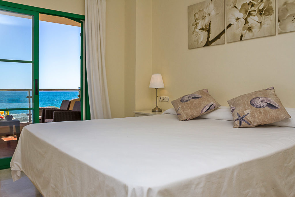 Image shows a large king size bed from which you can see the sea and beach and terrace